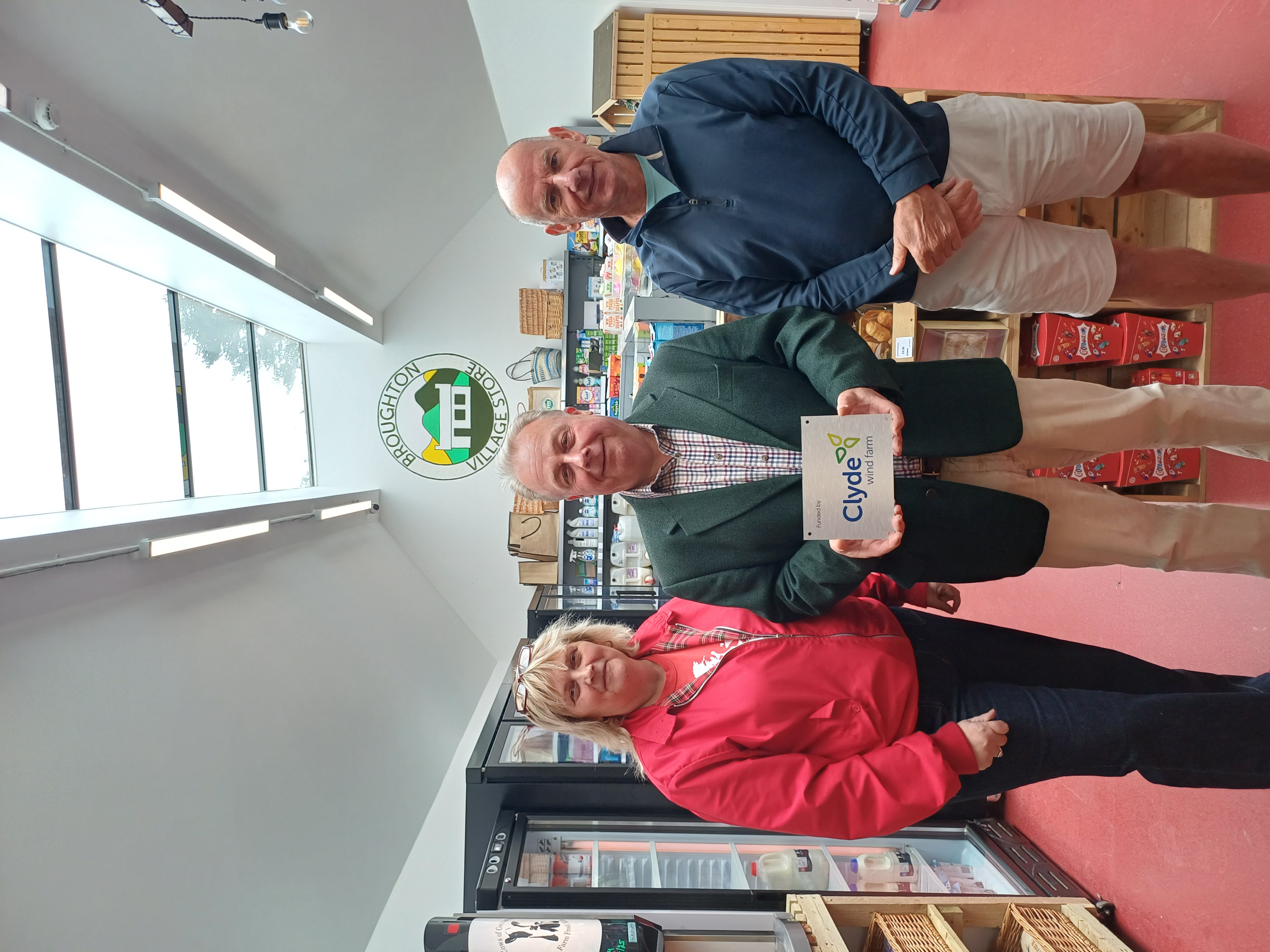 A woman and two men standing in the Broughton Village Store, holding a sign saying Clyde Wind Farm.