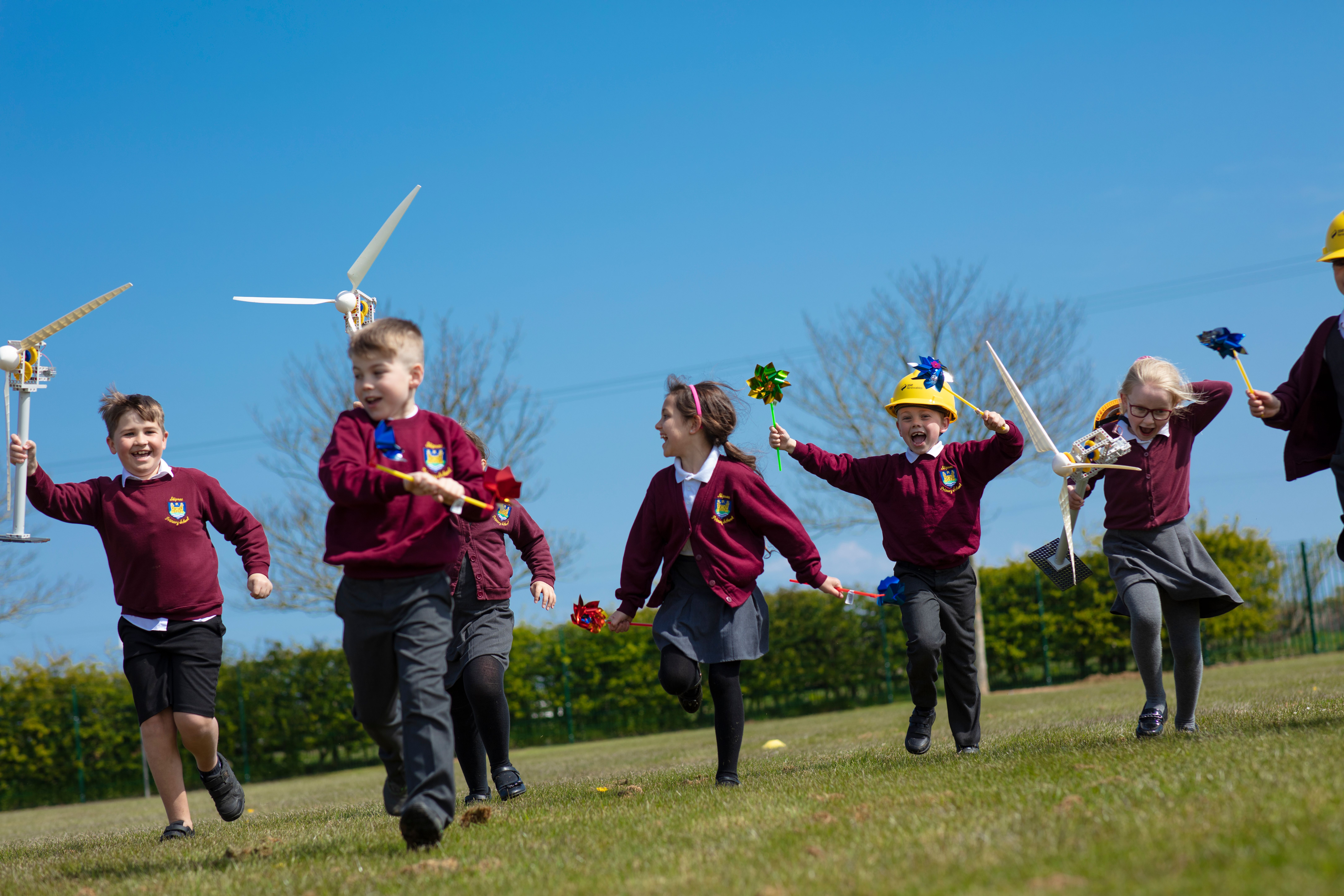Primary school children running happily across a field on a sunny day carrying toy windmills and models of wind turbines.