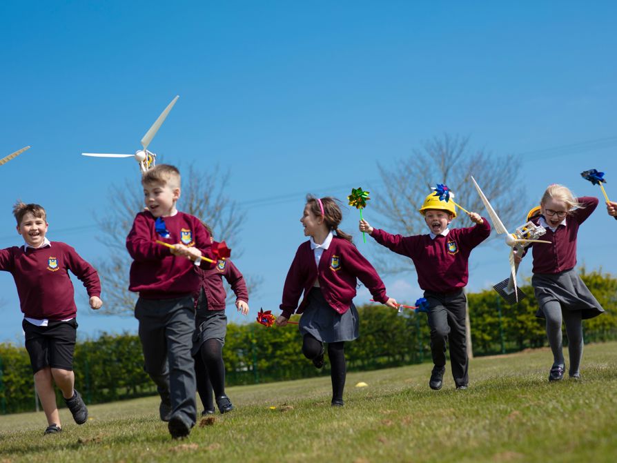 Primary school children running happily across a field on a sunny day carrying toy windmills and models of wind turbines.
